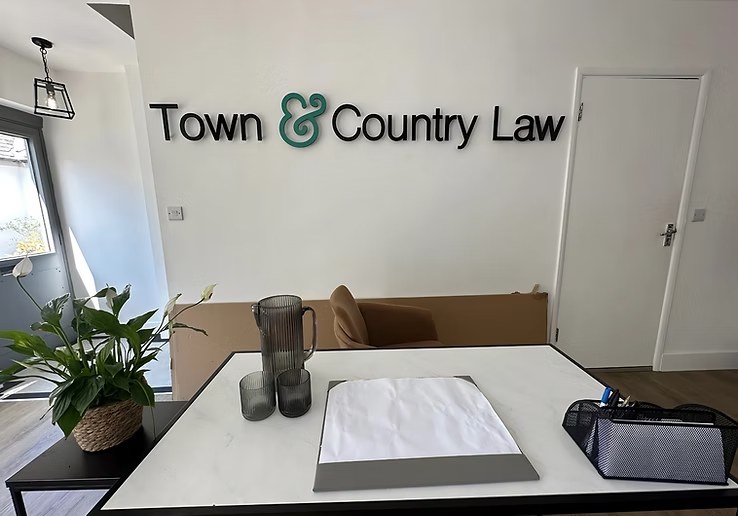 The reception wall at Town & Country Law Stamford: the wordmark mounted on white plaster, the green ampersand visible, a marble-topped table with peace lily and pendant lantern